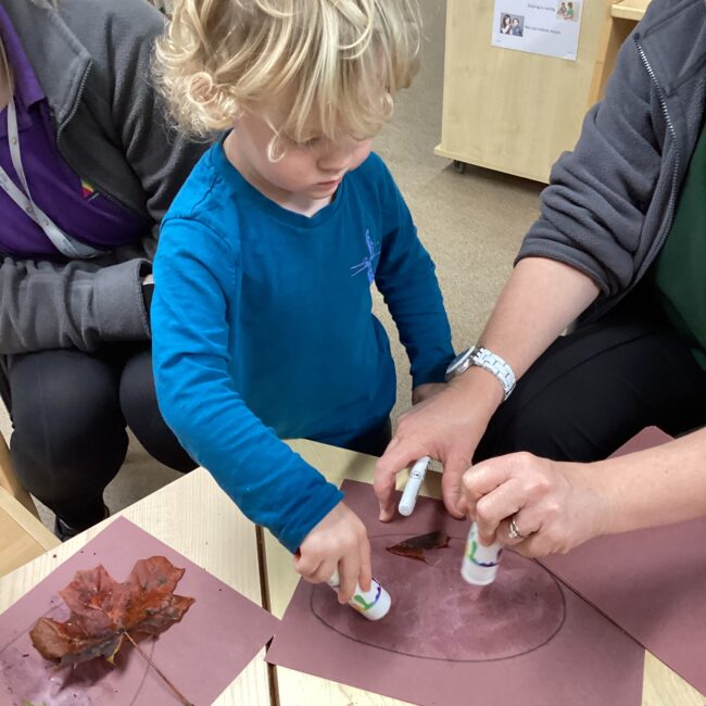 Infant child using glue sticks on paper at nursery.