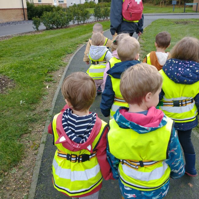 Infant children walking on path in hi-viz jackets led by nursery worker.