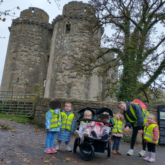 Nursery children in Hi-Viz jackets outside Nunney Castle with staff.