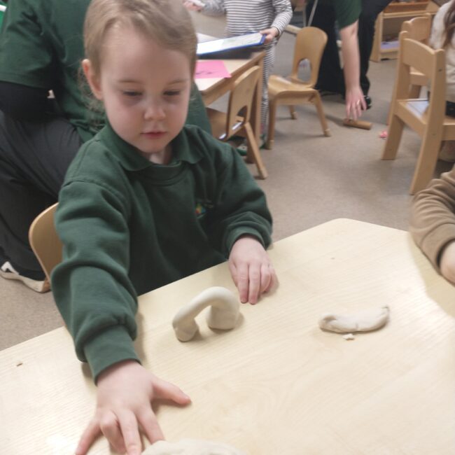 Infant child in green nursery uniform playing with clay at table.