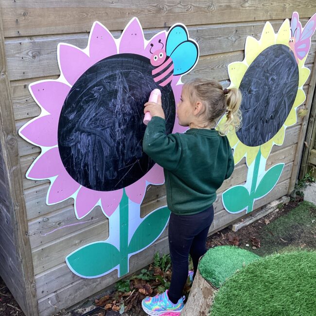 Infant girl using chalk on sunflower chalkboard outdoors at nursery.