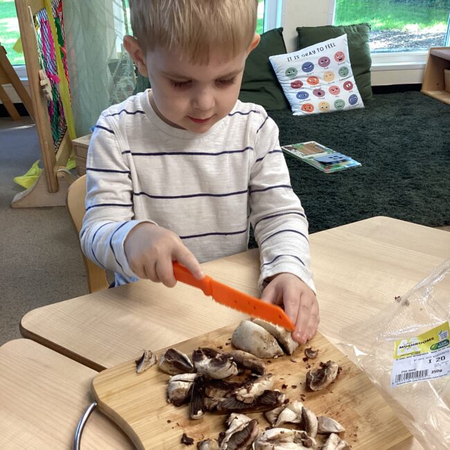 Infant boy chopping mushrooms on chopping board at nursery.